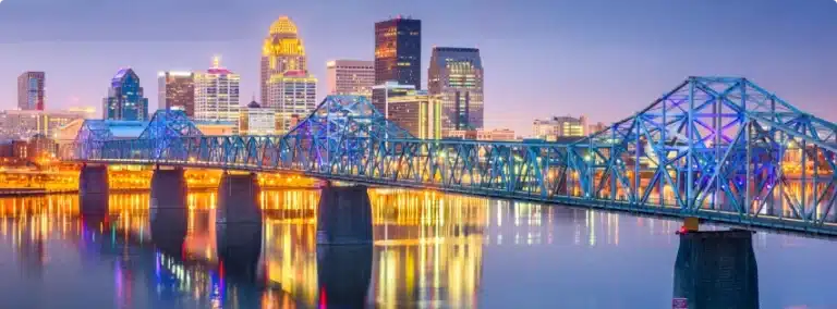 Kentucky city skyline at dusk with a brightly lit steel bridge spanning a calm river. The buildings are illuminated, reflecting vibrant colors on the water below.